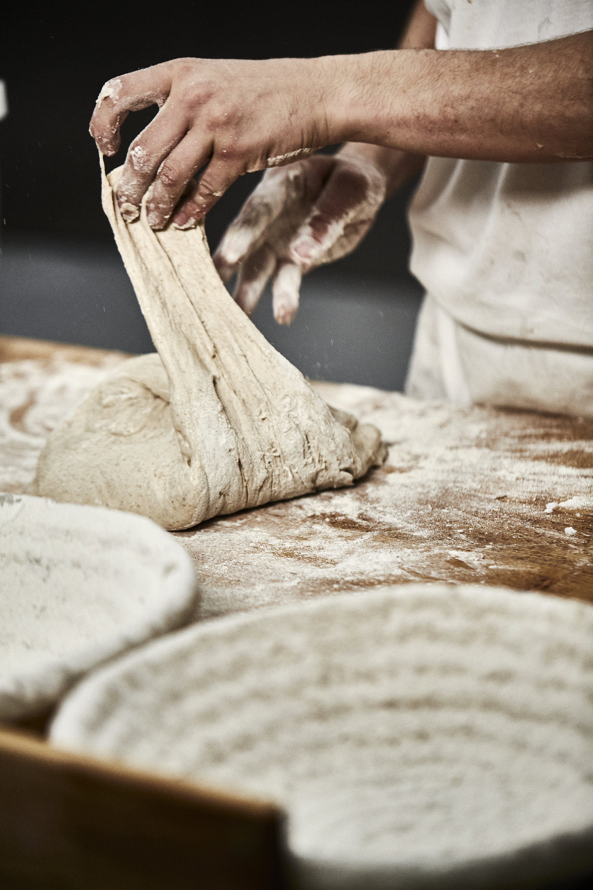 Bäckerhände verarbeiten einen Teig auf einer mehligen Arbeitsfläche. Im Vordergrund ist ein Korb zu sehen.