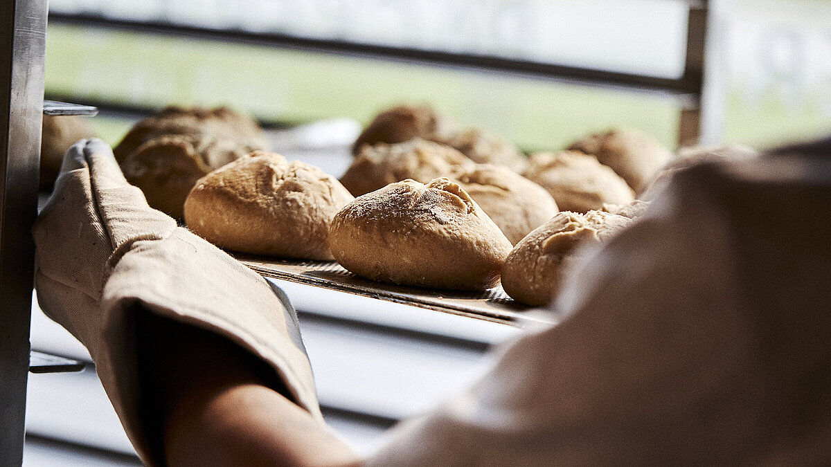 Blick über die linke Schulter eines Bäckers, der gerade Brötchen in eine Ablage schiebt
