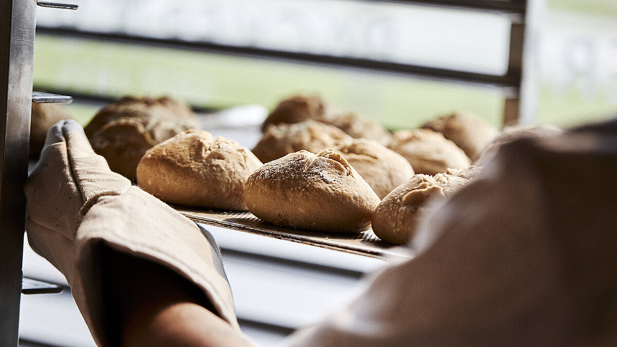 Blick über die linke Schulter eines Bäckers, der gerade Brötchen in eine Ablage schiebt