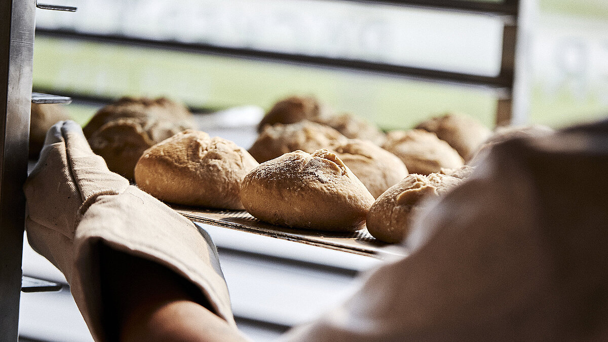 Blick über die linke Schulter eines Bäckers, der gerade Brötchen in eine Ablage schiebt
