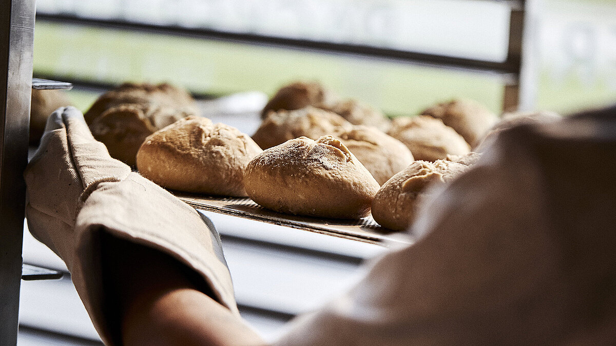 Blick über die linke Schulter eines Bäckers, der gerade Brötchen in eine Ablage schiebt