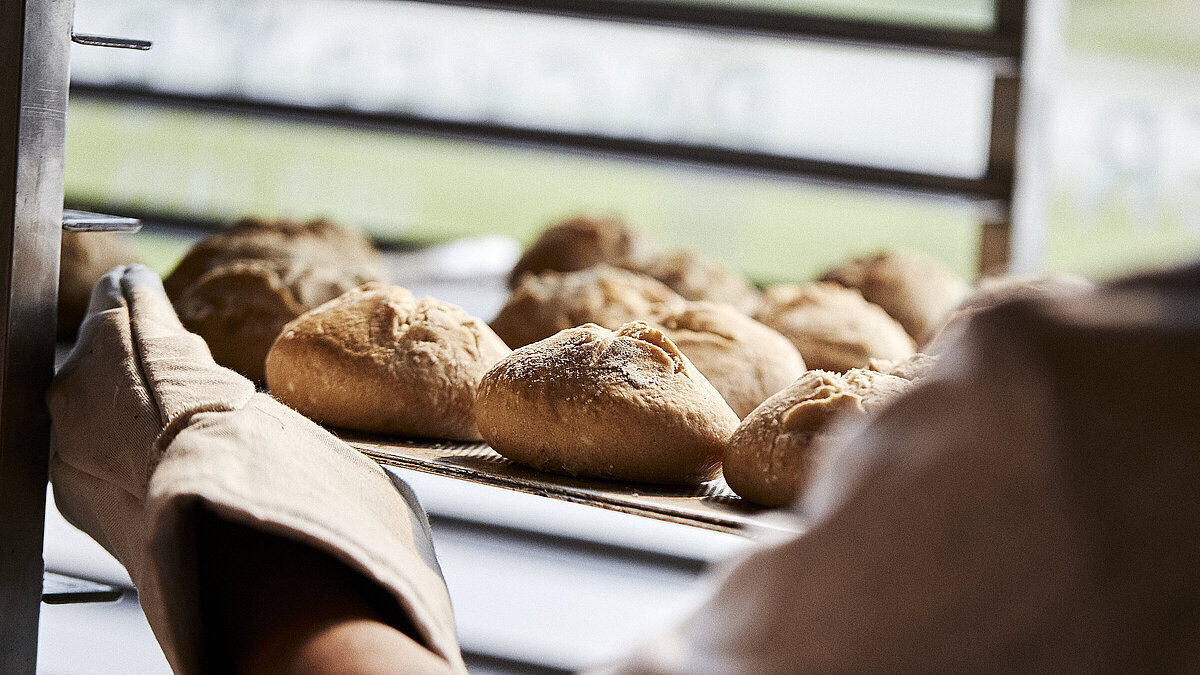Blick über die linke Schulter eines Bäckers, der gerade Brötchen in eine Ablage schiebt