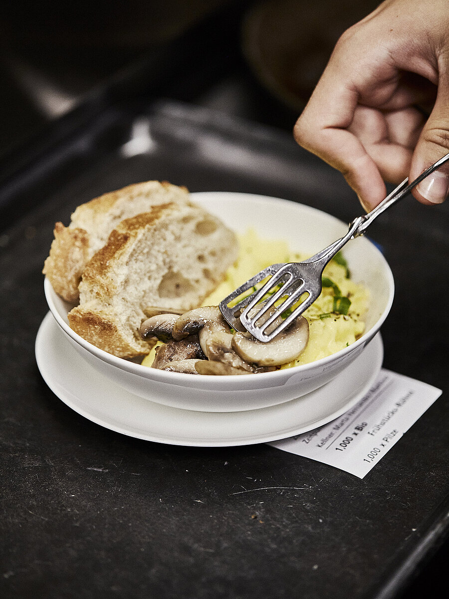 Person legt Champignons in einen Teller, in dem sich Rührei und zwei Scheiben weißes Brot befinden