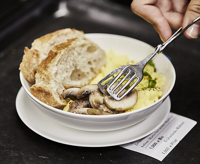 Person legt Champignons in einen Teller, in dem sich Rührei und zwei Scheiben weißes Brot befinden