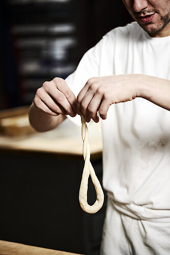 Bäcker formt eine Art Schleife aus Teig in der Luft. Jede Hand hält ein Teigende. 