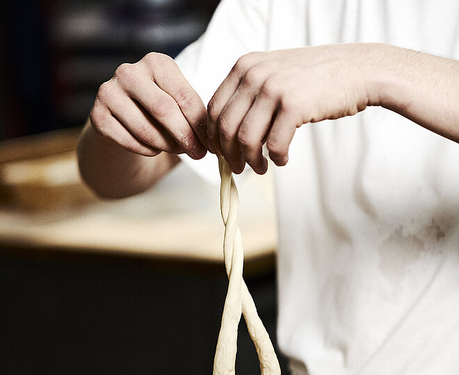 Bäcker formt eine Art Schleife aus Teig in der Luft. Jede Hand hält ein Teigende. 