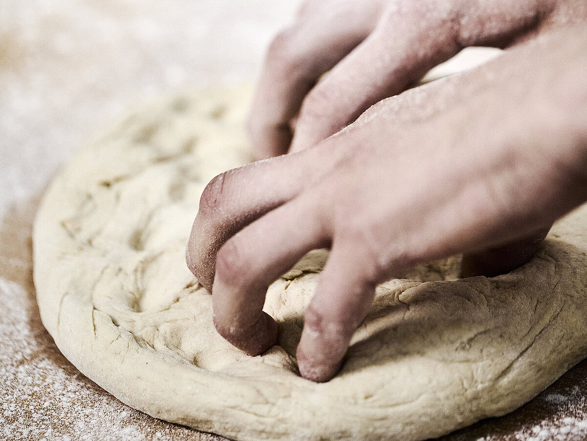 Bäckerhände bearbeiten einen flach ausgebreiteten Teig auf einer mehligen Arbeitsfläche. 