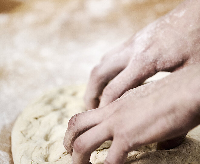 Bäckerhände bearbeiten einen flach ausgebreiteten Teig auf einer mehligen Arbeitsfläche. 