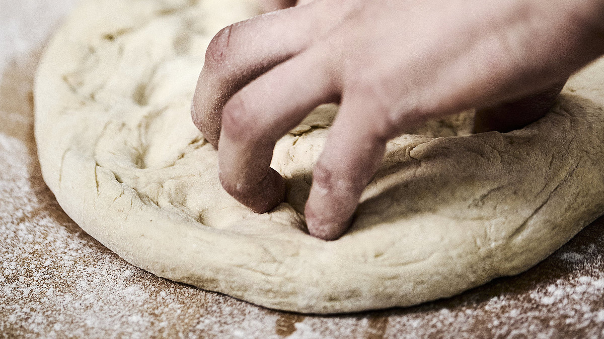 Bäckerhände bearbeiten einen flach ausgebreiteten Teig auf einer mehligen Arbeitsfläche. 