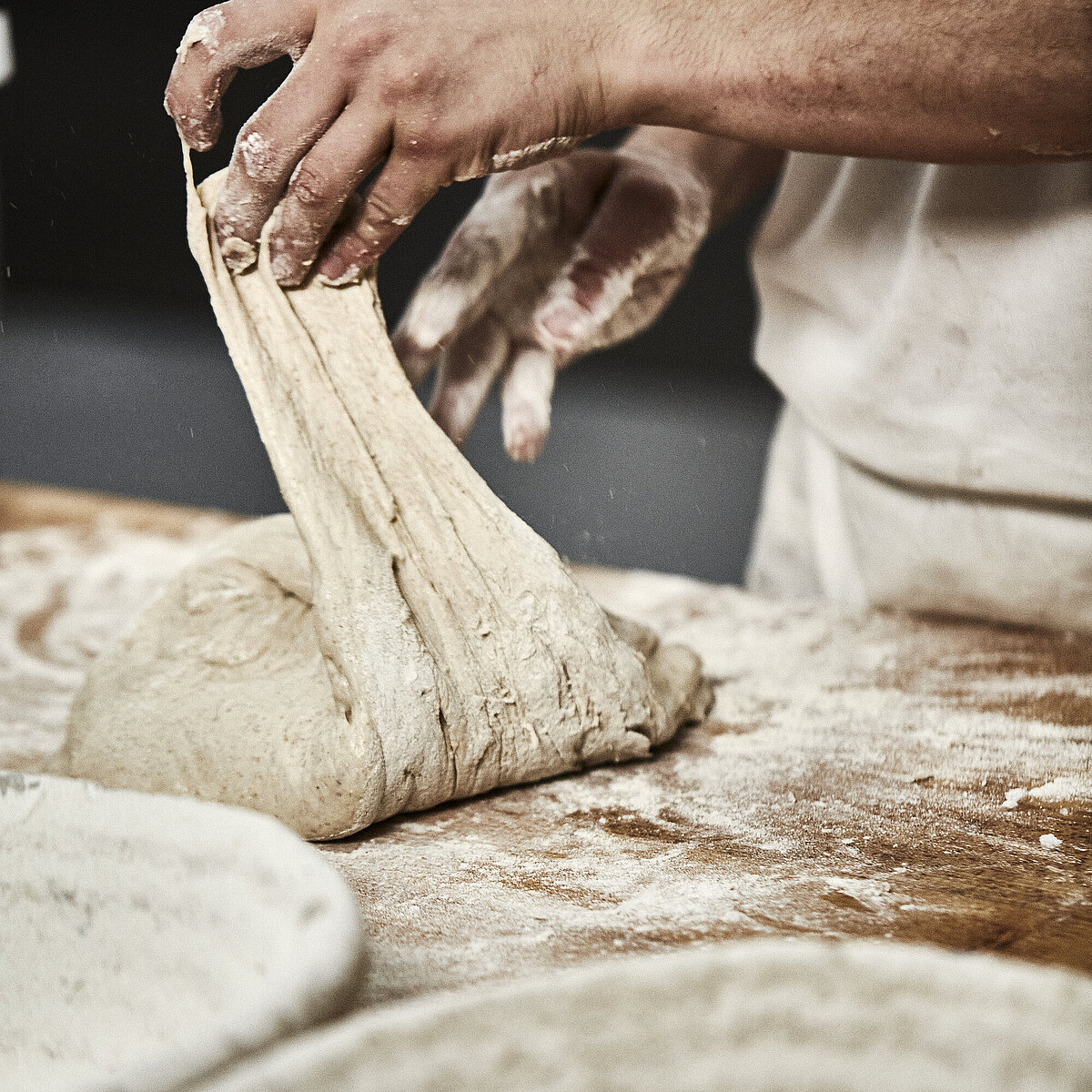Bäckerhände verarbeiten einen Teig auf einer mehligen Arbeitsfläche. Im Vordergrund ist ein Korb zu sehen.