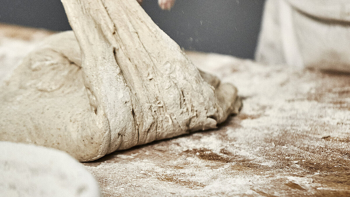 Bäckerhände verarbeiten einen Teig auf einer mehligen Arbeitsfläche. Im Vordergrund ist ein Korb zu sehen.