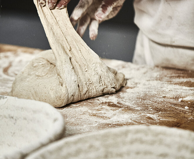 Bäckerhände verarbeiten einen Teig auf einer mehligen Arbeitsfläche. Im Vordergrund ist ein Korb zu sehen.