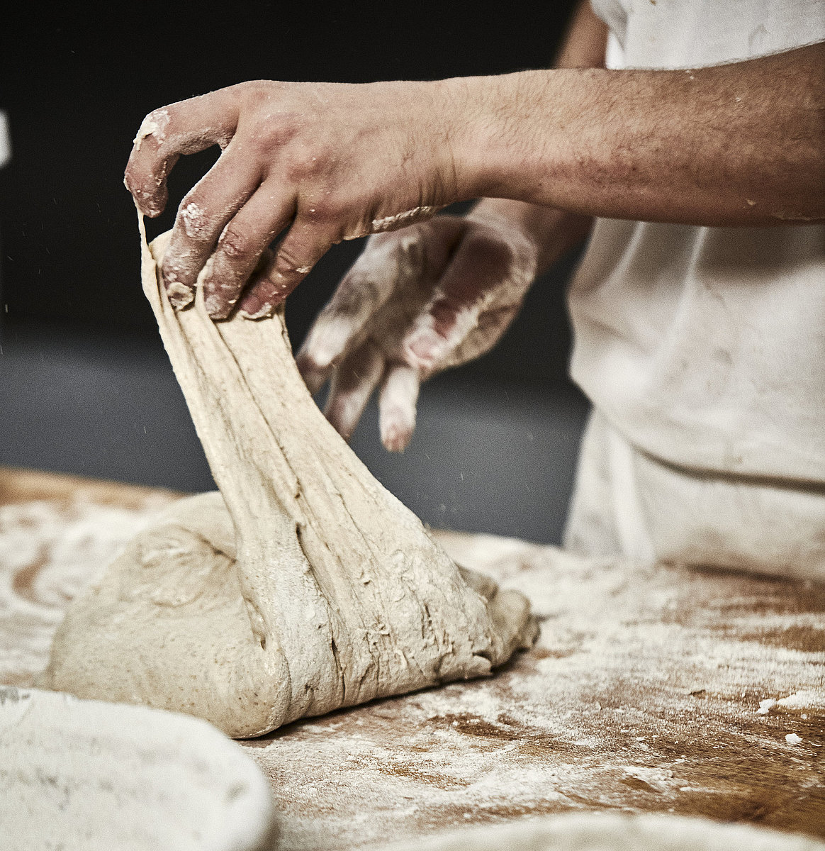 Bäckerhände verarbeiten einen Teig auf einer mehligen Arbeitsfläche. Im Vordergrund ist ein Korb zu sehen.