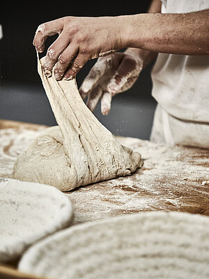 Bäckerhände verarbeiten einen Teig auf einer mehligen Arbeitsfläche. Im Vordergrund ist ein Korb zu sehen.