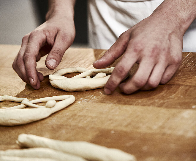 Bäckerhände formen eine Laugenbreze aus Teig. Im Vordergrund sind Teigrollen zu sehen.
