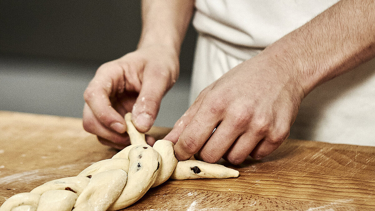 Bäckerhände flechten einen Zopf aus Teig auf einer mehligen Arbeitsfläche.