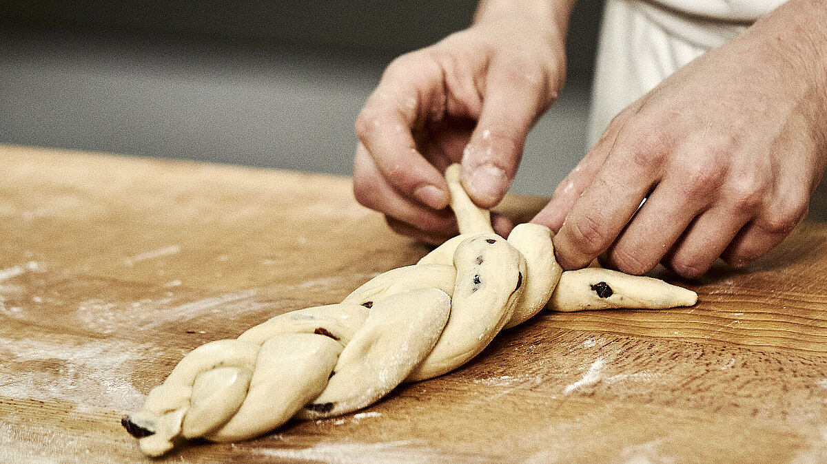 Bäckerhände flechten einen Zopf aus Teig auf einer mehligen Arbeitsfläche.