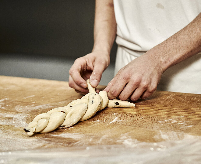 Bäckerhände flechten einen Zopf aus Teig auf einer mehligen Arbeitsfläche.