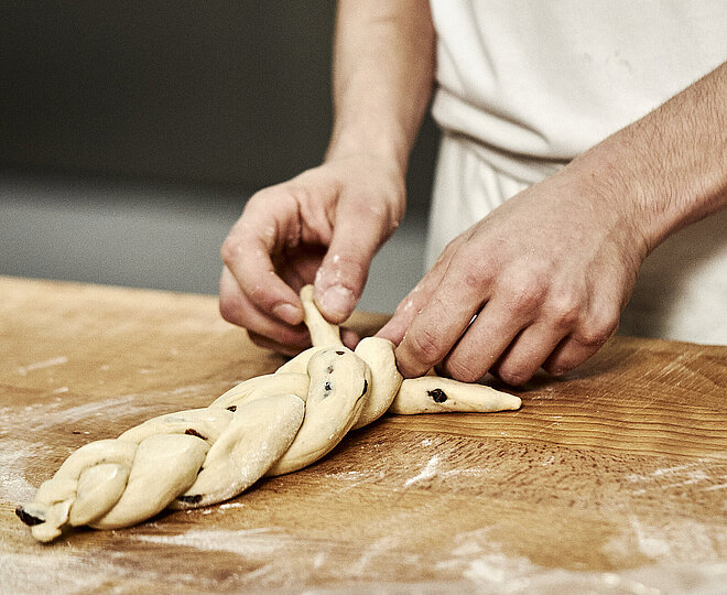 Bäckerhände flechten einen Zopf aus Teig auf einer mehligen Arbeitsfläche.