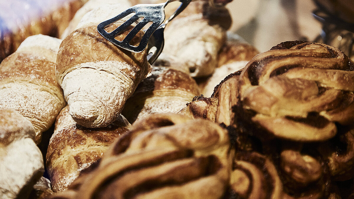 Person greift mit einer metallischen Gebäckzange nach eine Croissant, dass inmitten mehrerer süßen Backwaren liegt. 