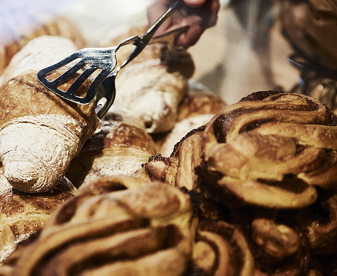 Person greift mit einer metallischen Gebäckzange nach eine Croissant, dass inmitten mehrerer süßen Backwaren liegt. 
