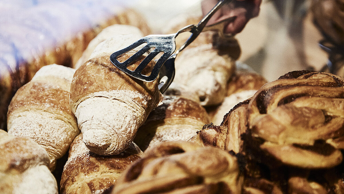 Person greift mit einer metallischen Gebäckzange nach eine Croissant, dass inmitten mehrerer süßen Backwaren liegt. 