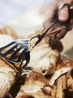 Person greift mit einer metallischen Gebäckzange nach eine Croissant, dass inmitten mehrerer süßen Backwaren liegt. 