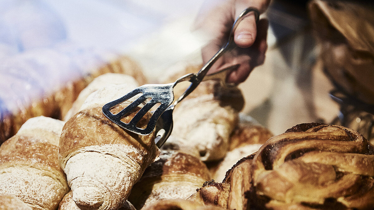 Person greift mit einer metallischen Gebäckzange nach eine Croissant, dass inmitten mehrerer süßen Backwaren liegt. 