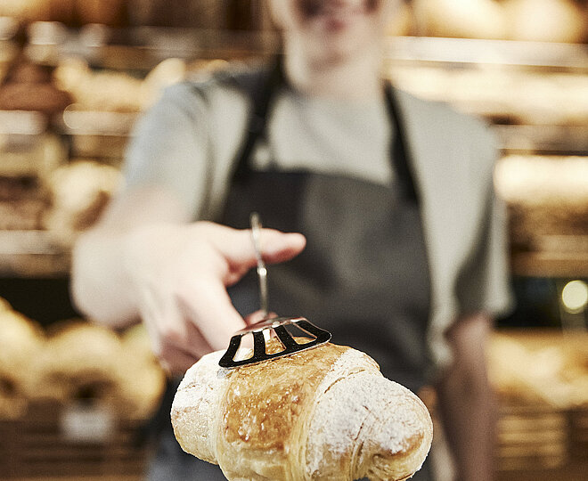 Weibliche Person mit grauer Kochschürze reicht ein Croissant in die Kamera, das Croissant ist scharf, die Person ist unscharf.
