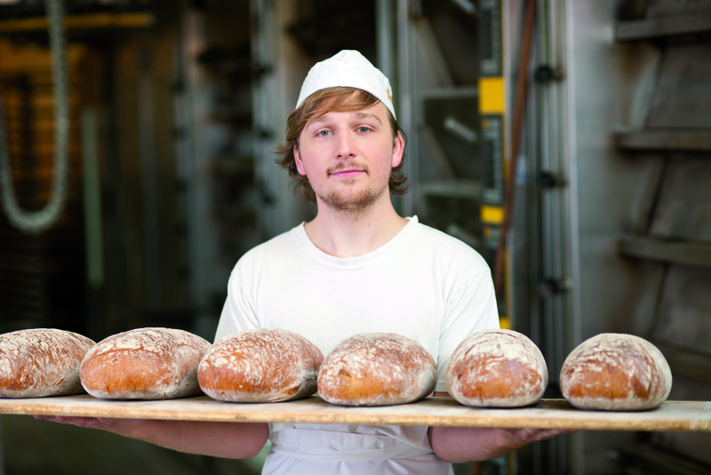 Auszubildender trägt Holzbrett mit frisch gebackenen Broten