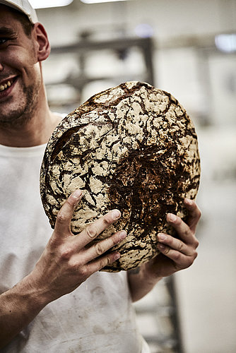 Bäcler hält ein großes, dunkles Brot in beiden Händen. Die Person ist seitlich angeschnitten.