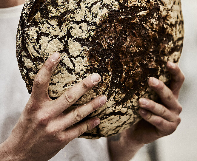 Bäcler hält ein großes, dunkles Brot in beiden Händen. Die Person ist seitlich angeschnitten.