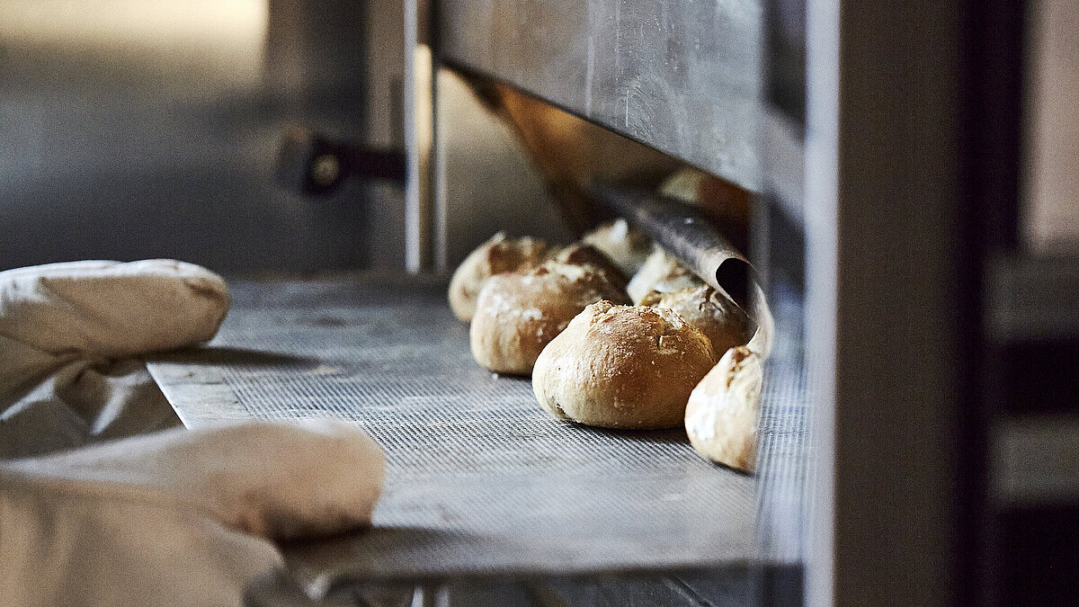Bäcker mit großen Handschuhen schiebt ein Backblech mit Brötchen in den Ofen.