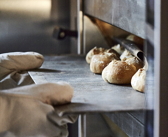 Bäcker mit großen Handschuhen schiebt ein Backblech mit Brötchen in den Ofen.