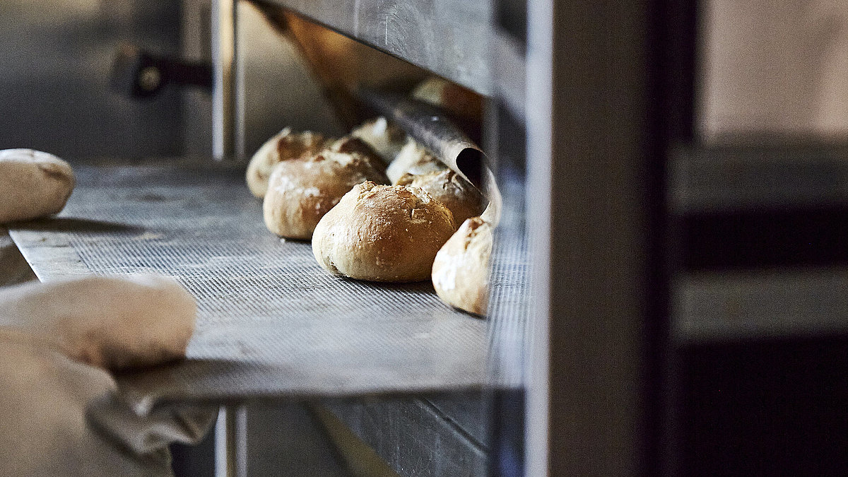 Bäcker mit großen Handschuhen schiebt ein Backblech mit Brötchen in den Ofen.