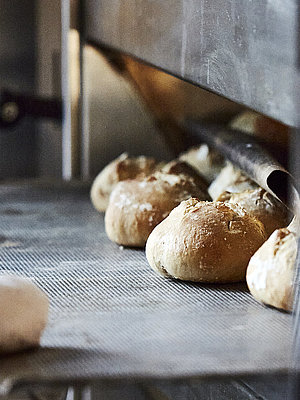 Bäcker mit großen Handschuhen schiebt ein Backblech mit Brötchen in den Ofen.