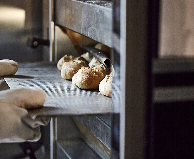 Bäcker mit großen Handschuhen schiebt ein Backblech mit Brötchen in den Ofen.