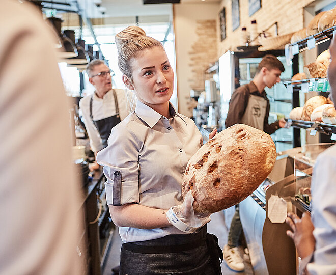 Bäckereifachverkäuferin spricht mit einer Kollegin und hält dabei ein Brot
