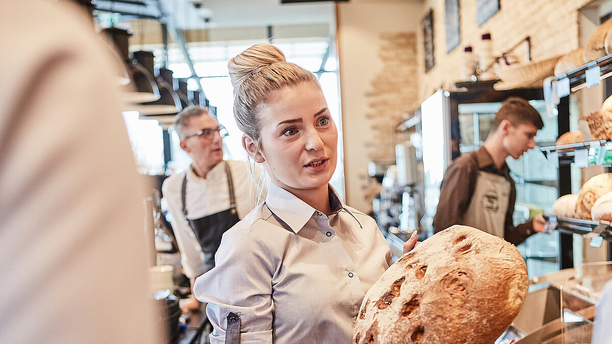 Bäckereifachverkäuferin spricht mit einer Kollegin und hält dabei ein Brot