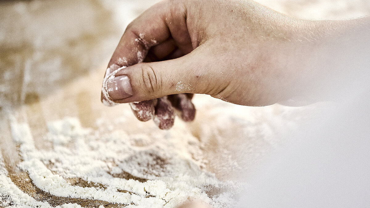 Detailaufnahme einer Bäckerhand, die Mehl auf einer Arbeitsfläche verteilt.