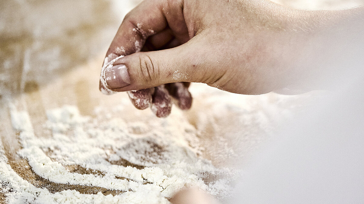 Detailaufnahme einer Bäckerhand, die Mehl auf einer Arbeitsfläche verteilt.