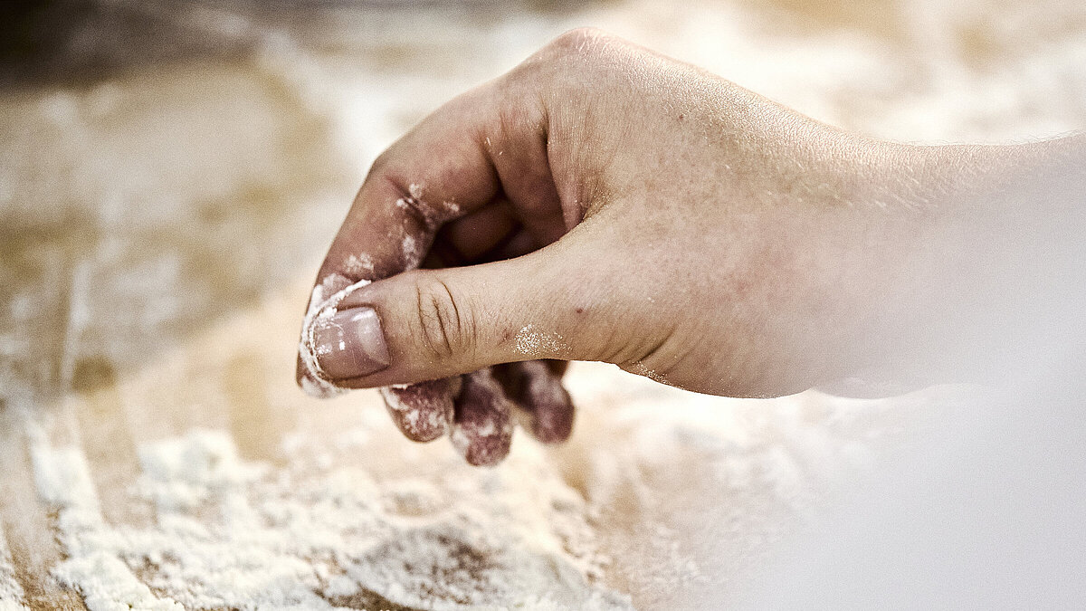 Detailaufnahme einer Bäckerhand, die Mehl auf einer Arbeitsfläche verteilt.